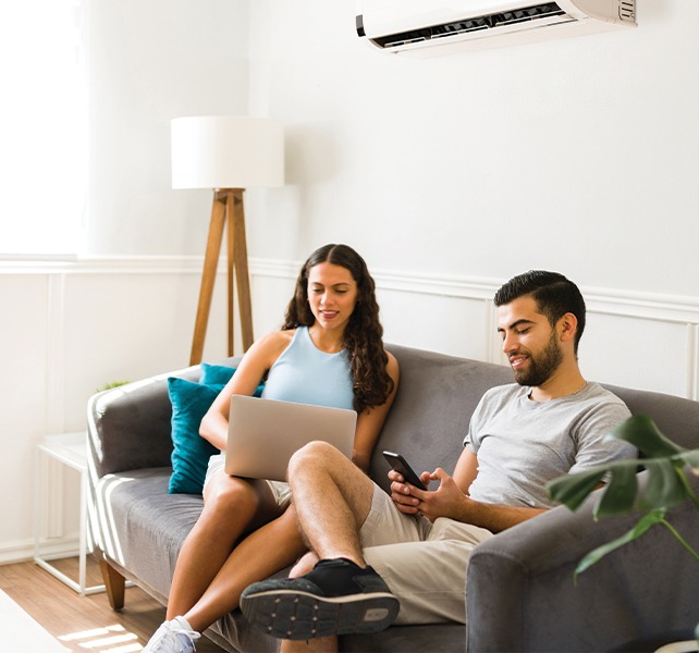 Un couple détendu dans un salon moderne, avec un homme utilisant son téléphone et une femme travaillant sur un ordinateur portable, sous un climatiseur mural. Cette image illustre le confort et la technologie dans un espace de vie, idéal pour accompagner un article sur les thermostats intelligents.